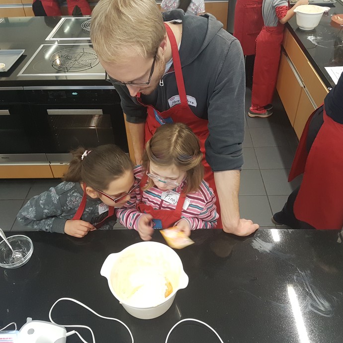 Zwei Kinder schauen in eine Teigschüssel. Ein Lehrer schaut ihnen über die Schulter. (vergrößerte Bildansicht wird geöffnet)