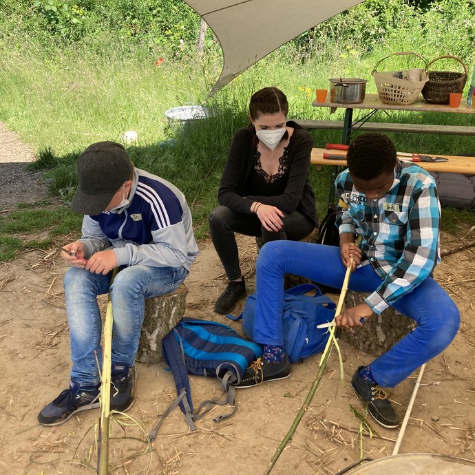 Zwei Jungen schnitzen an langen Stöcken. Eine Frau sitzt dabei und schaut zu. (vergrößerte Bildansicht wird geöffnet)