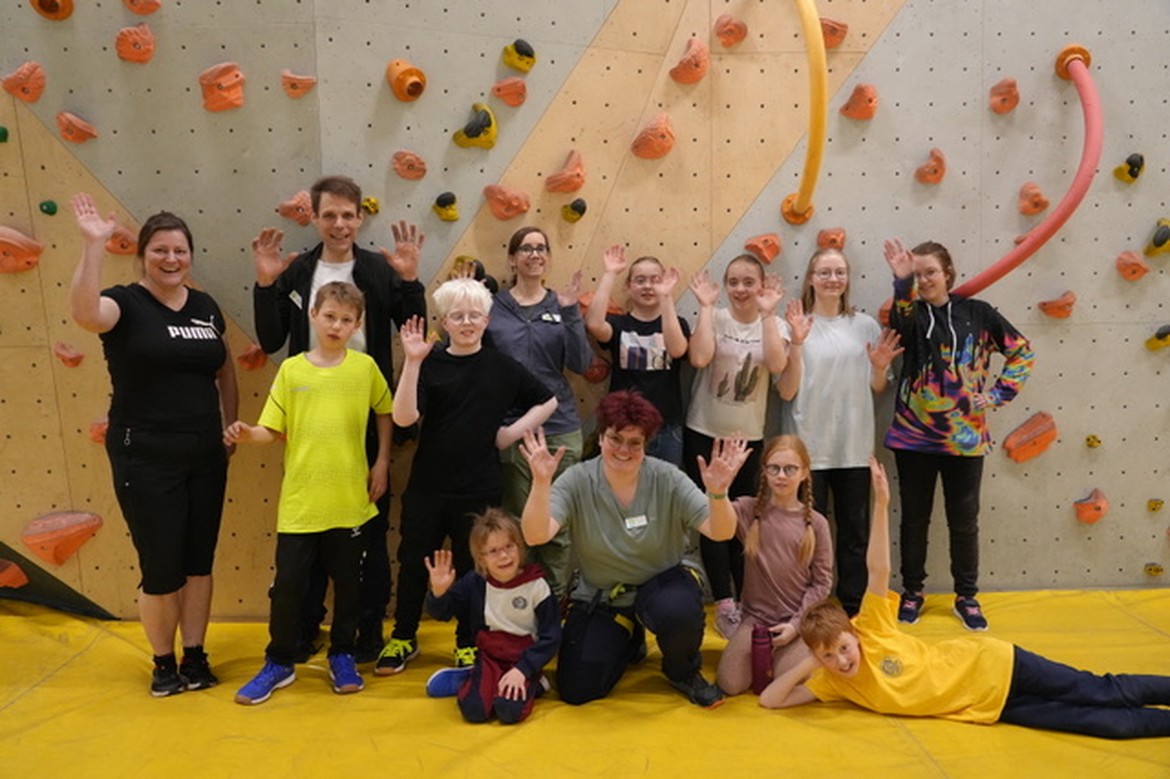 Mehrere Schülerinnen und Schüler und zwei Erwachsene stehen vor einer Kletterwand und winken.