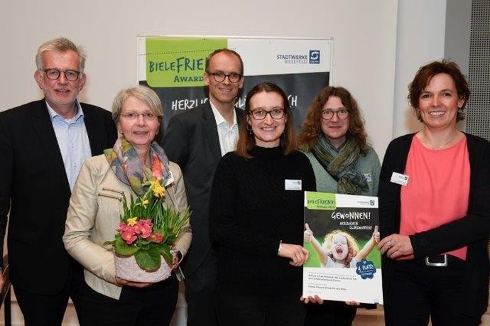 Mehrere Erwachsene stehen vor einem Schild mit der Aufschrift "Bielefeld Awards". Eine Lehrerin hält einen Blumenstrauß, eine andere eine Urkunde.