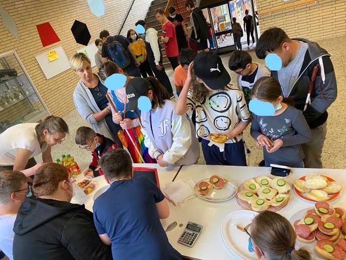 Mehrere Schüler*innen stehen vor einem Tisch, auf dem Brötchen und Getränke liegen, und kaufen ein.