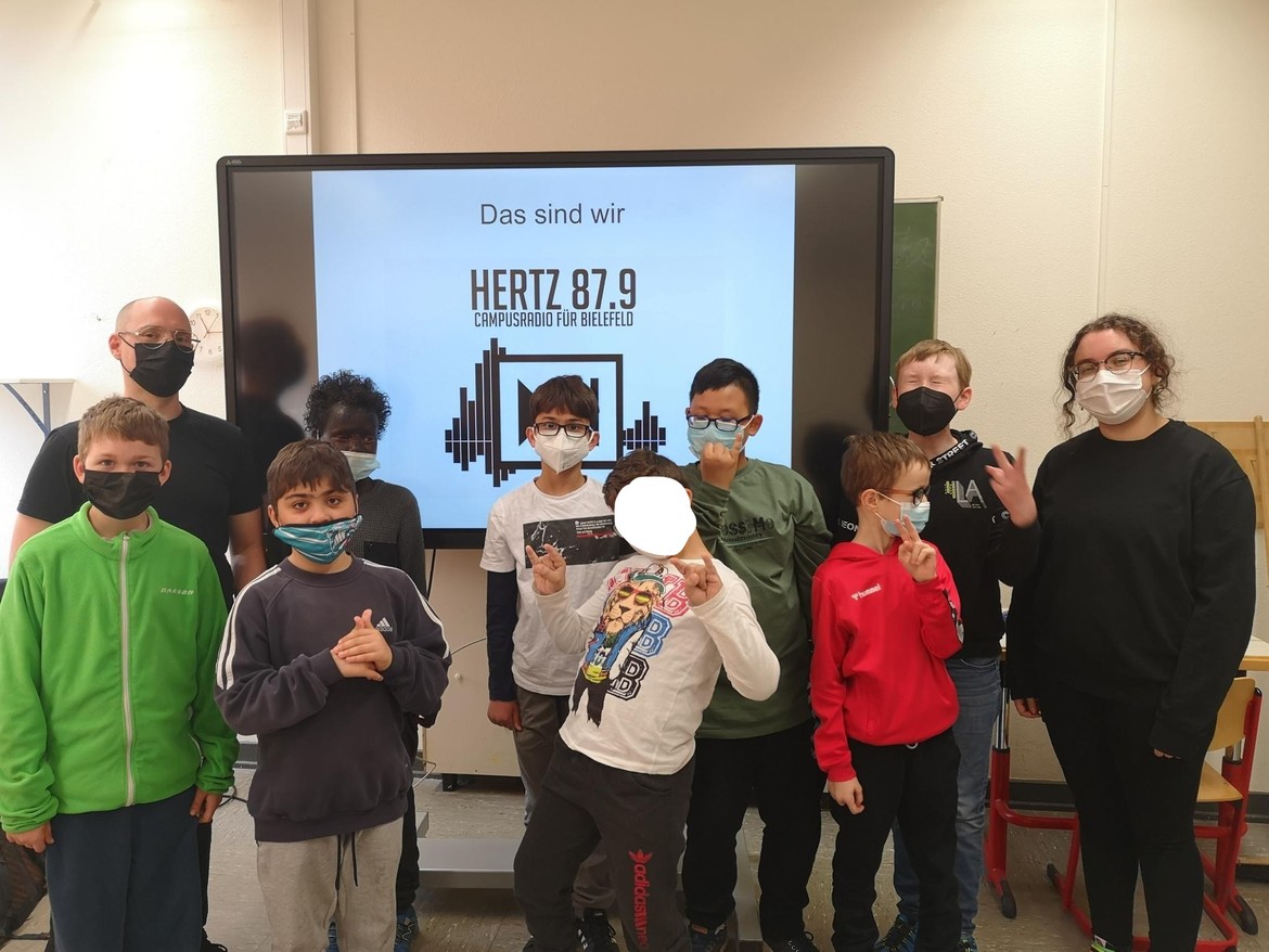Einige Kinder und zwei Erwachsene mit Masken stehen vor einem Smartboard mit der Aufschrift "Hertz 87.9".