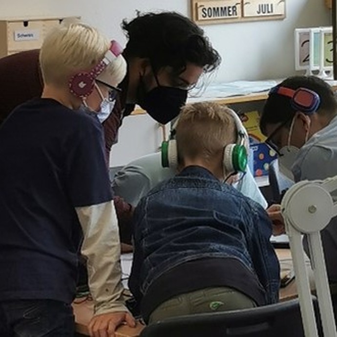 Drei Kinder mit Kopfhörern hören einem Mann zu. Alle tragen Masken. (vergrößerte Bildansicht wird geöffnet)