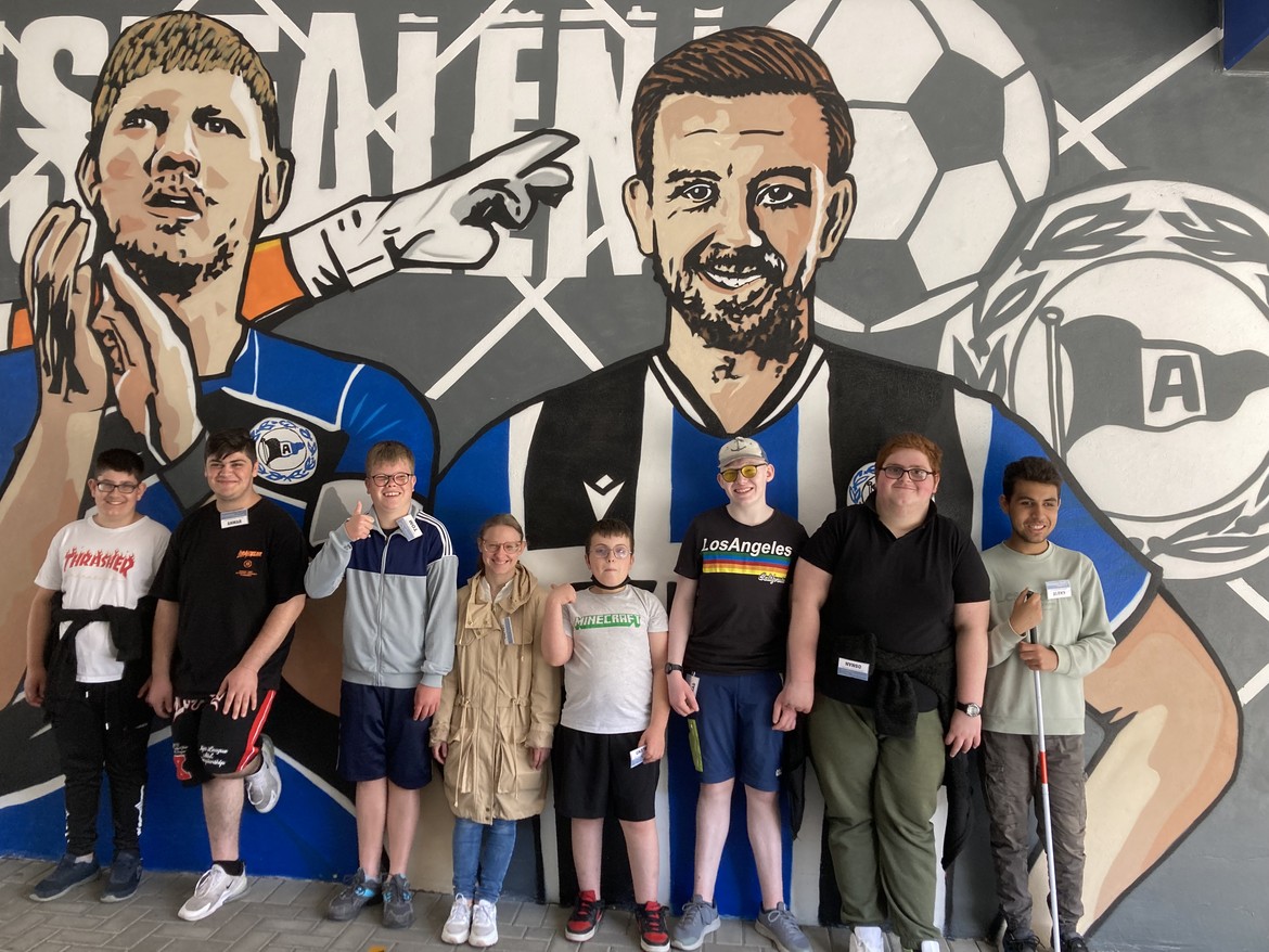 Die Schüler*innen der Klasse 6-10 stehen vor einer Wand mit großen gemalten Bildern der Arminia-Spieler.