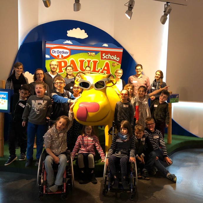 Eine Gruppe von Kindern und Lehrer*innen steht um eine große gelbe Kuh (Werbemaskottchen von Paula-Pudding). (vergrößerte Bildansicht wird geöffnet)