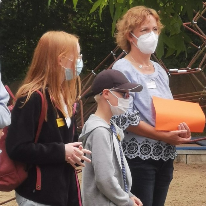 Eine Lehrerin steht auf dem Schulhof und erzählt etwas. Zwei Kinder hören zu. Alle tragen Masken. (vergrößerte Bildansicht wird geöffnet)