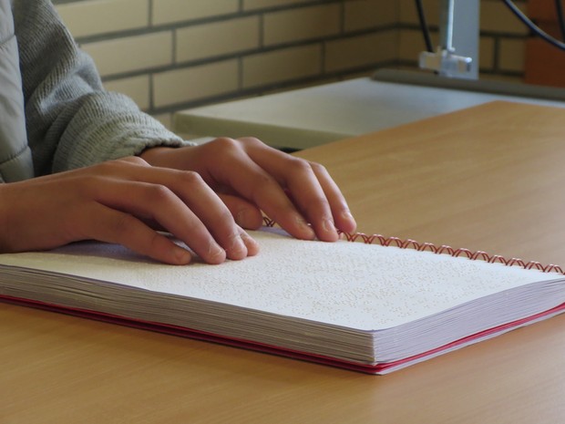 Ein Punktschrift-Text liegt auf dem Tisch. Der Leser oder die Leserin hat die Hände darauf in Leseposition platziert. Ein Punktschrift-Text liegt auf dem Tisch. Der Leser oder die Leserin hat die Hände darauf in Leseposition platziert.