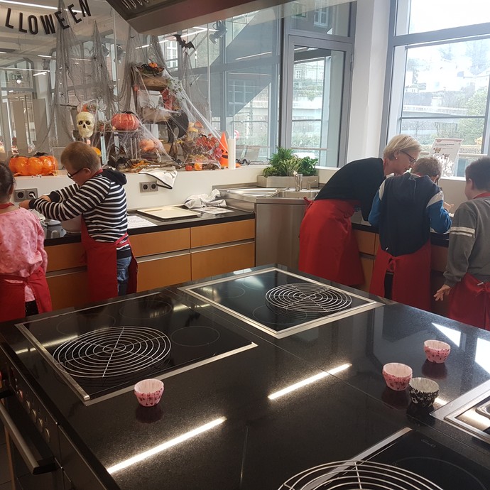Kinder stehen mit Schürzen in einer Küche. Eine Lehrerin hilft einem Kind beim Teigkneten. (vergrößerte Bildansicht wird geöffnet)