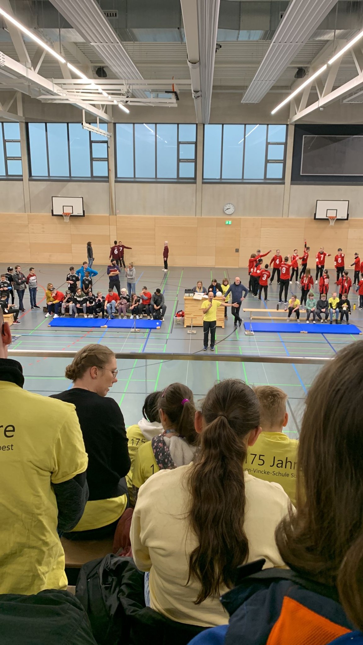 In einer Sporthalle sitzen viele Kinder und Jugendliche, einige laufen über das Feld.