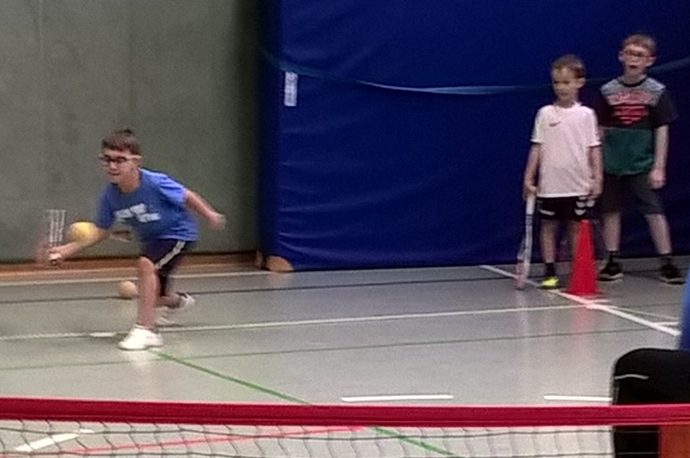 Ein Schüler rennt und schwingt einen Tennisschläger. Zwei weitere Jungen stehen vor einer dicken Matte und schauen ihm zu.
